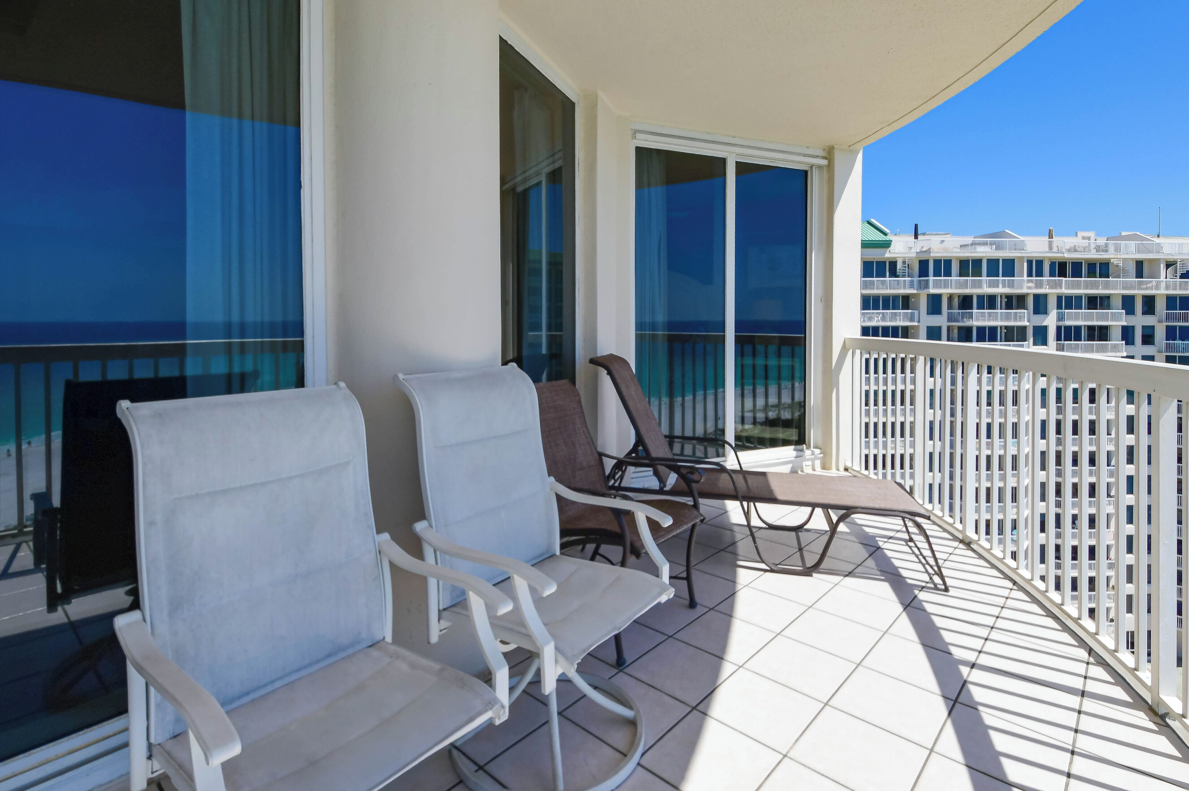 A balcony with two white chairs and a table, large windows, and a view of a building complex.