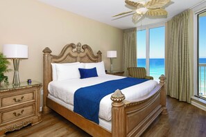 Una cama grande con cabecera de madera, dos mesitas de noche con lámparas, una mesita con un jarrón, y vista al océano a través de amplios ventanales.
