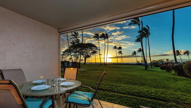 A dining area with a table set for four, overlooking a well-manicured lawn and palm trees, with a sunset view through large windows. - Leinaala Oceanfront #104  Ocean Front, A/C, Oceanfront  views, Remodeled! (Kihei)