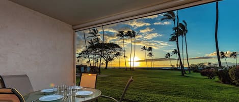 Un coin repas avec une table dressée pour quatre personnes, donnant sur une pelouse bien entretenue et des palmiers, et offrant une vue sur le coucher de soleil à travers de grandes fenêtres.