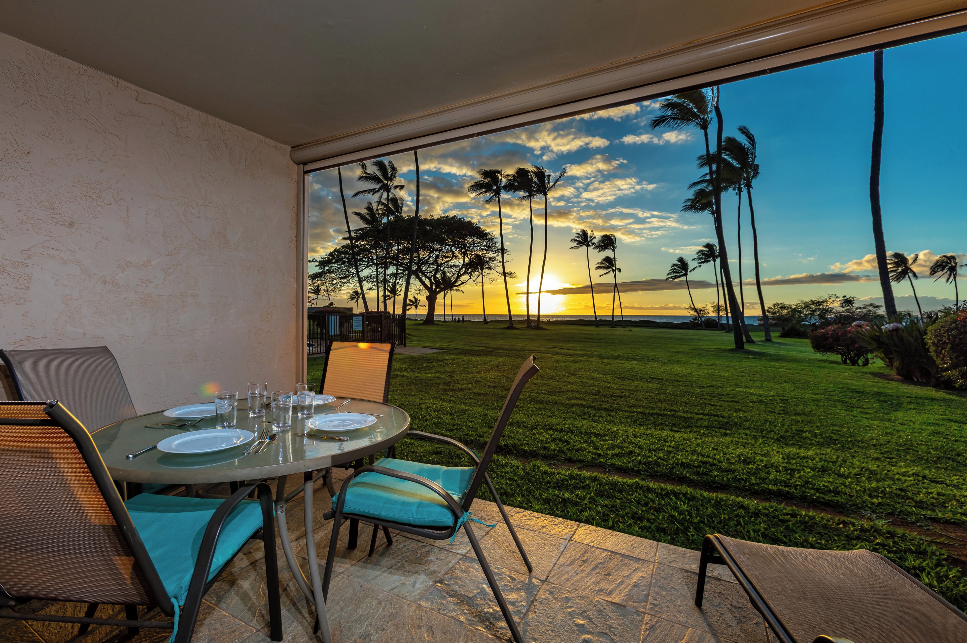 A dining area with a table set for four, overlooking a well-manicured lawn and palm trees, with a sunset view through large windows.