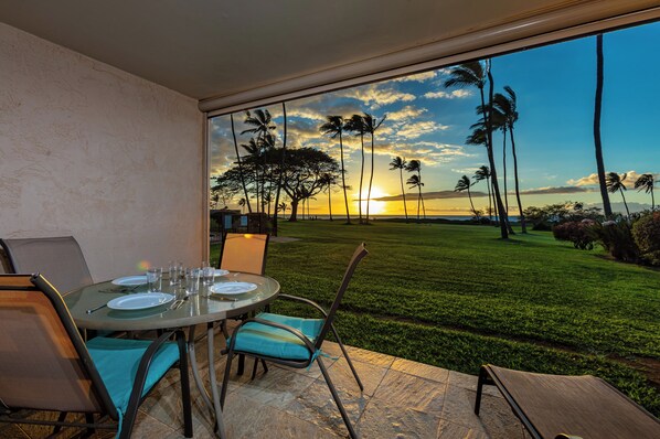 A dining area with a table set for four, overlooking a well-manicured lawn and palm trees, with a sunset view through large windows.