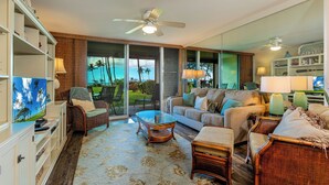 A spacious living room with a large TV, wicker furniture, and a view of palm trees through the sliding glass doors. - Leinaala Oceanfront #104  Ocean Front, A/C, Oceanfront  views, Remodeled! (Kihei)