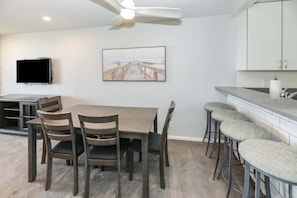 A dining area with a table and chairs, a television mounted on the wall, and a kitchen area with a counter and stools. - Modern Kitchen w/ Ocean & Harbor View- Upper Cottage, G32  North Coast Village (Oceanside)