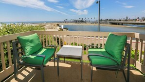 A balcony with two green cushioned chairs and a small table, overlooking a beach and a marina. - Modern Kitchen w/ Ocean & Harbor View- Upper Cottage, G32  North Coast Village (Oceanside)
