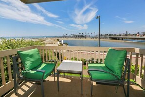A balcony with two green cushioned chairs and a small table, overlooking a beach and a marina. - Modern Kitchen w/ Ocean & Harbor View- Upper Cottage, G32  North Coast Village (Oceanside)