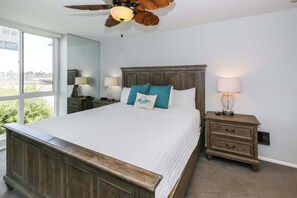 A bedroom with a large bed, wooden headboard, bedside tables with lamps, a ceiling fan, and a view through a window. - Modern Kitchen w/ Ocean & Harbor View- Upper Cottage, G32  North Coast Village (Oceanside)