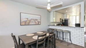 A dining area with a table and chairs, a ceiling fan, a kitchen area with a counter, and a large mirror reflecting the kitchen. - Modern Kitchen w/ Ocean & Harbor View- Upper Cottage, G32  North Coast Village (Oceanside)