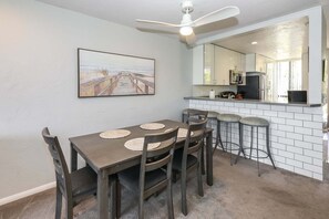 A dining area with a table and chairs, a ceiling fan, a kitchen area with a counter, and a large mirror reflecting the kitchen. - Modern Kitchen w/ Ocean & Harbor View- Upper Cottage, G32  North Coast Village (Oceanside)