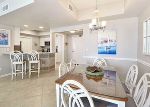 A dining area with a glass-top table, white chairs, a chandelier, and a painting of a sailboat on the wall. - Fresh, fun, & bright beachfront condo w/private balcony! (Destin)
