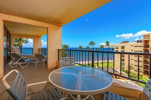 Ein Balkon mit Glastisch und Stühlen, der Blick auf einen Strand mit Palmen und einem wolkenlosen, blauen Himmel bietet.