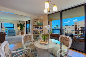 A modern living room with a glass table, wicker chairs, a bookshelf, and a balcony with a view. - Oceanview Penthouse Condo - Spacious & Serene Sanctuary - Sugar Beach #PH12 (Kihei)