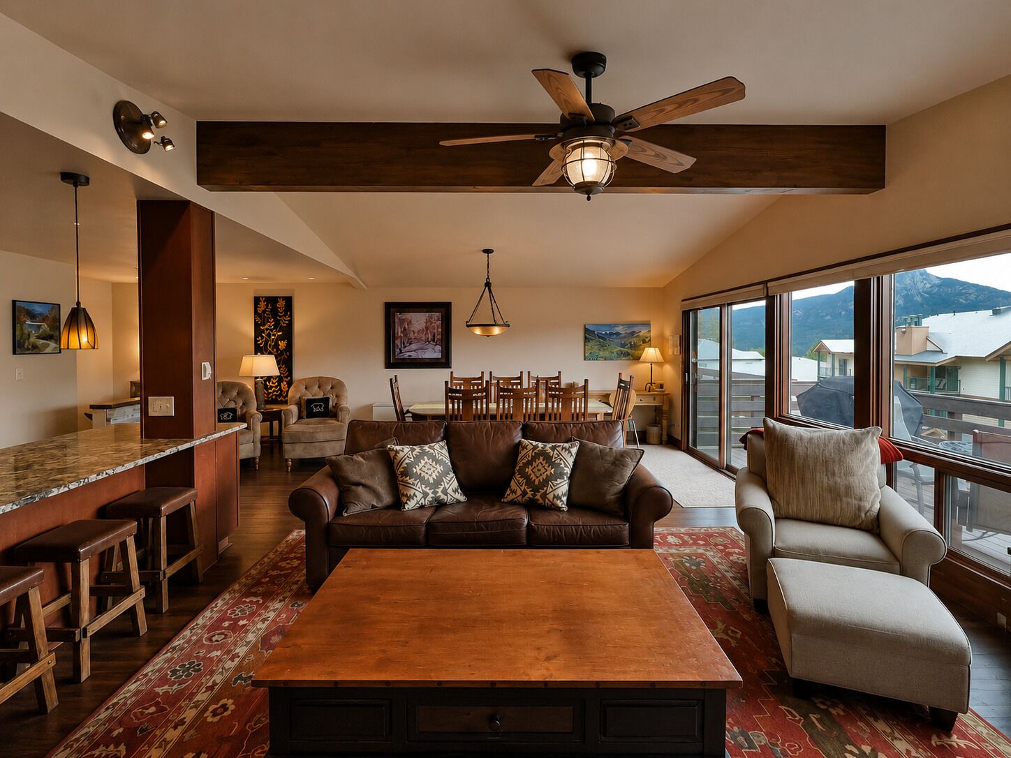 A spacious living room with a large wooden coffee table, a sofa, and a chair. There is a ceiling fan, a dining area with chairs, and a view through large windows.