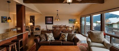A spacious living room with a large wooden coffee table, a sofa, and a chair. There is a ceiling fan, a dining area with chairs, and a view through large windows.