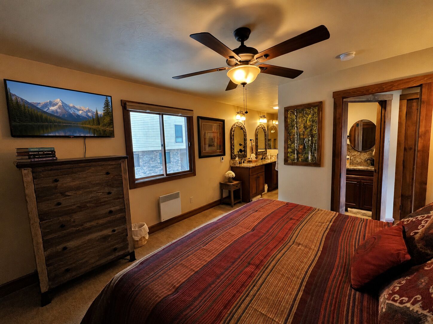 A bedroom with a bed, a ceiling fan, a large window, a dresser, a bathroom with a sink and mirror, and a painting on the wall.