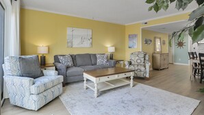 Una sala de estar con sofá, sillón y mesa de centro, un comedor con sillas y una mesa, y una cocina al fondo.