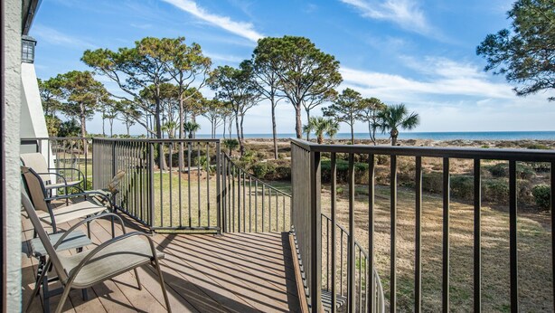 Una terraza de madera con barandas de metal, mobiliario exterior y vistas a la playa y árboles.