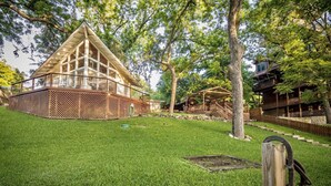 Una cabaña de madera con terraza, rodeada por un césped cuidado y árboles.