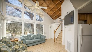 Una sala de estar amplia con un gran ventanal, ventilador de techo, sofá y una zona de cocina con refrigerador.