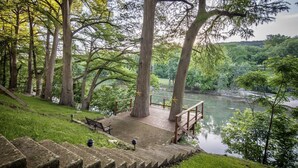 Un parque con una pasarela de madera que da a un río, rodeado de árboles y una zona verde con un banco.