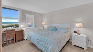 A bedroom with a large bed, two bedside tables with lamps, a dresser, a desk with a chair, and a window with a view of the beach. - Beachfront Condo Offers Fun and Sun At Bargain Price! PET FRIENDLY! (Fort Myers Beach)