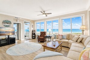 A spacious living room with a large TV, comfortable seating, a ceiling fan, and a view of the beach.