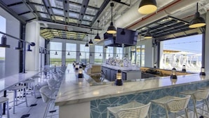 A modern bar area with a long counter, bar stools, pendant lighting, and large windows offering views of the outdoors. - Golf Cart! Pet-friendly! Linkside Village 435! (Miramar Beach)