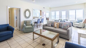 A spacious living room with a large window, a dining area, and a kitchen visible through an open door. - Gulf front! Pet Friendly! Beach House 30A Unit #2 - Gulf Front, Dune Allen Beach (Santa Rosa Beach)
