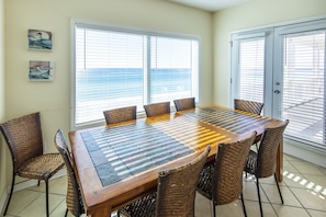 A dining area with a large wooden table and wicker chairs, large windows with blinds, and artwork on the wall. - Gulf front! Pet Friendly! Beach House 30A Unit #2 - Gulf Front, Dune Allen Beach (Santa Rosa Beach)