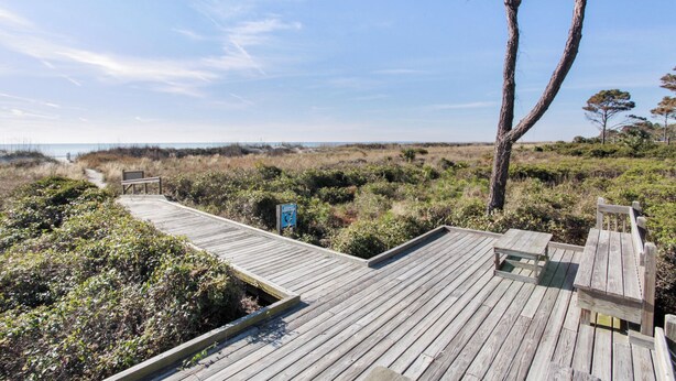 Une promenade en bois menant à une plage, avec un panneau indicateur et un arbre.