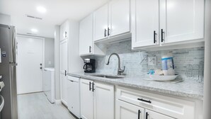 Une cuisine moderne avec des meubles blancs, un réfrigérateur en acier inoxydable, une machine à café et un évier avec un dosseret en carreaux bleus.