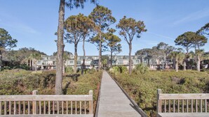 Une promenade en bois mène à un ensemble de bâtiments modernes, entourés de grands arbres et d’une végétation dense.