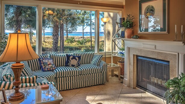 Una habitación luminosa con un sofá a rayas, una mesa con una lámpara, una chimenea y vistas a la playa.