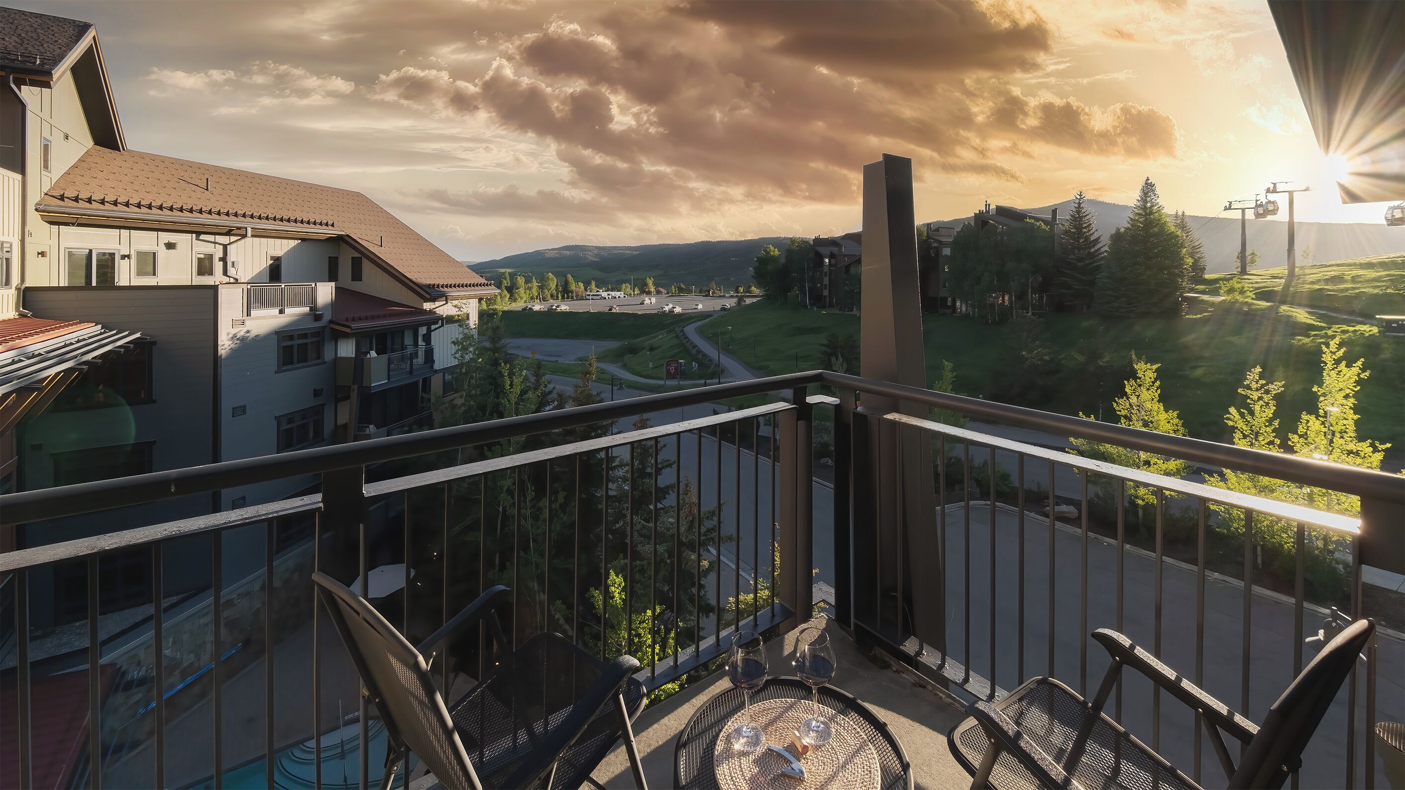 Varanda com vista para uma estrada e edifícios, piscina, mesa com cadeiras e copos de vinho.