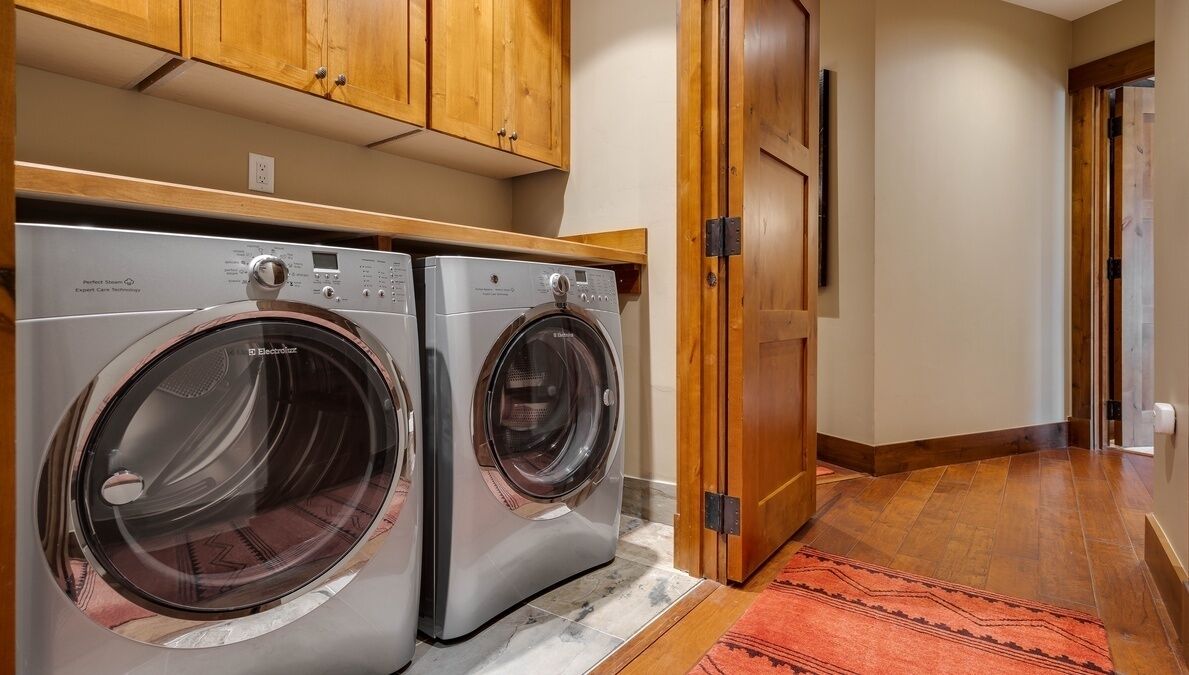 A laundry room with two front-loading washing machines, wooden cabinets above, and a red rug on the floor.