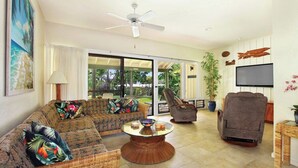A living room with a sofa, armchair, and a coffee table. There is a ceiling fan, a TV, and a sliding glass door leading to an outdoor area.