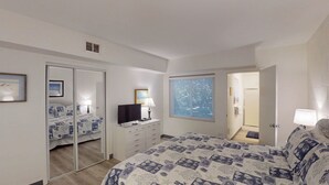 A bedroom with a bed, a dresser, a television, a window with blinds, a mirror, and a door leading to a bathroom.