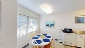 A compact kitchen with white appliances, a dining table with blue placemats, and a painting of boats on the wall.