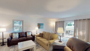 A living room with a brown leather sofa, a beige sofa, a small white table, and a lamp. There is a window with blinds and a view of the outside.
