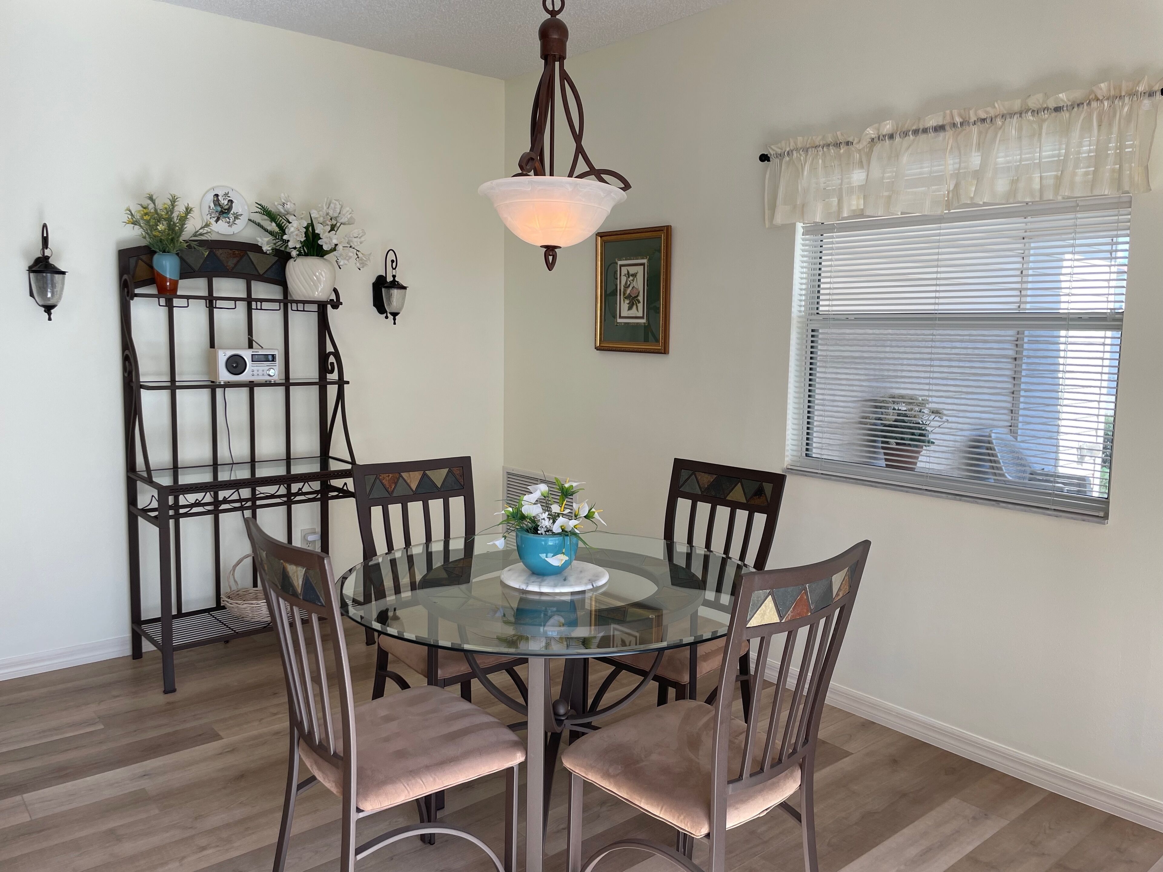 A dining area with a glass table surrounded by chairs, a decorative metal shelf with plants, a hanging light fixture, and a window with blinds.