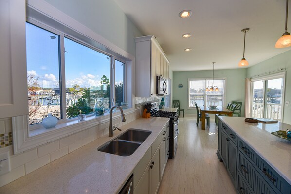 A kitchen with a large window, a sink, cabinets, and a dining area with a table and chairs. - Beautiful home-waterfront, beach, pools, family fun & pets! (Long Neck)