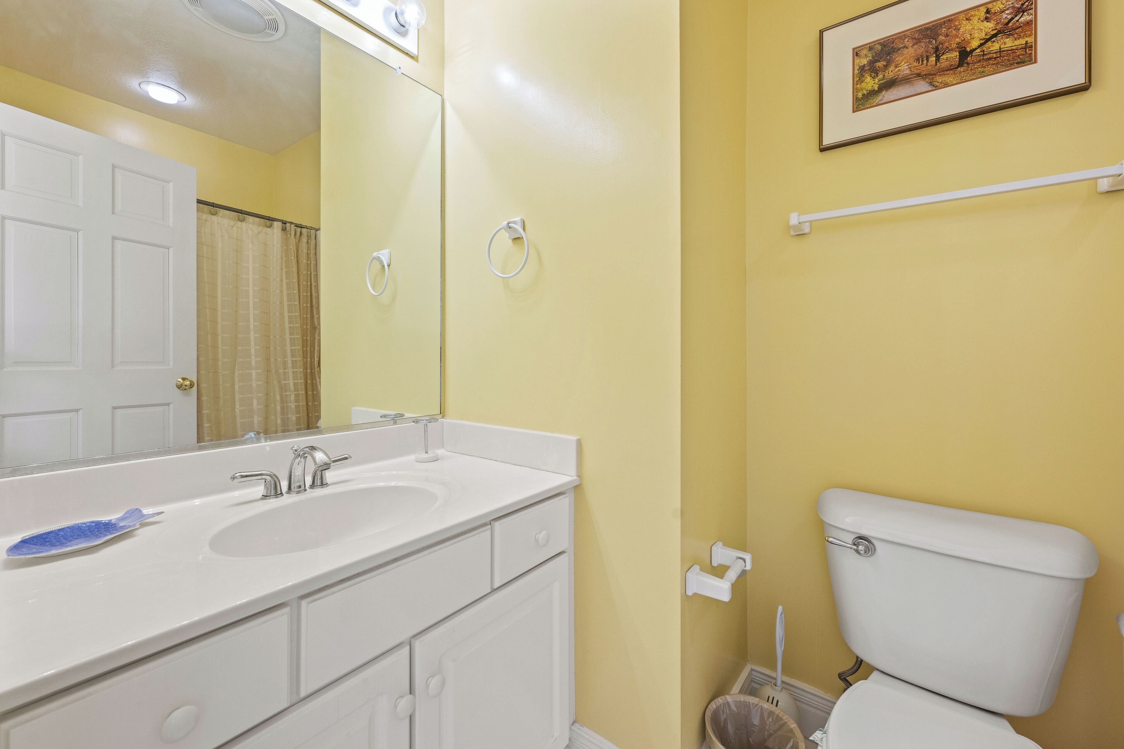 A bathroom with a white sink, mirror, and toilet. There is a framed picture on the wall and a towel rack.