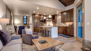 A modern kitchen with wooden cabinets, a central island, and pendant lighting. There is a dining area with a table and chairs, and a living room with a sofa and coffee table.