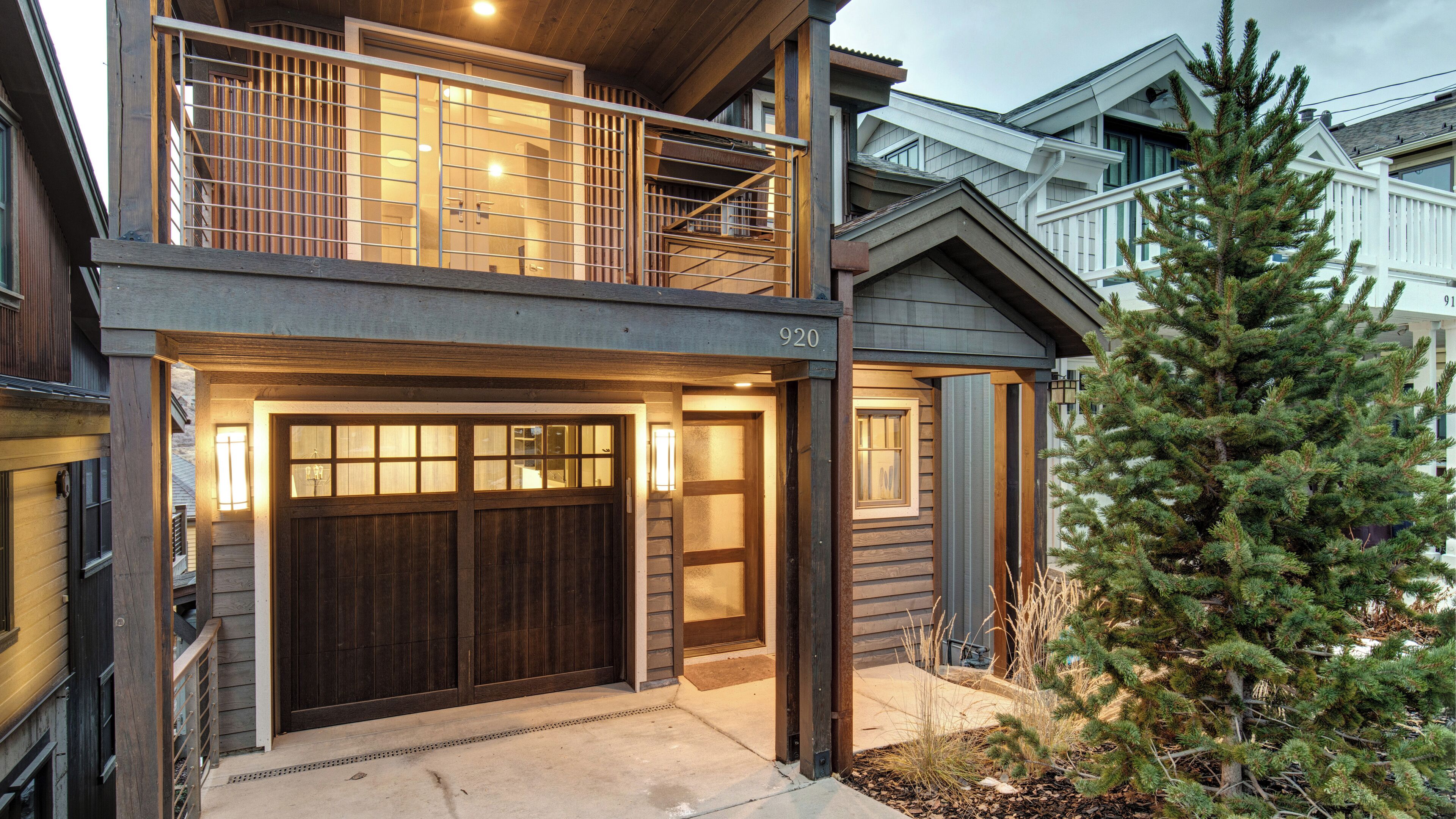 Ein zweistöckiges Haus mit einer dunklen Holztür zur Garage, einem Balkon mit Metallgeländer und einem großen immergrünen Baum im Vorgarten.