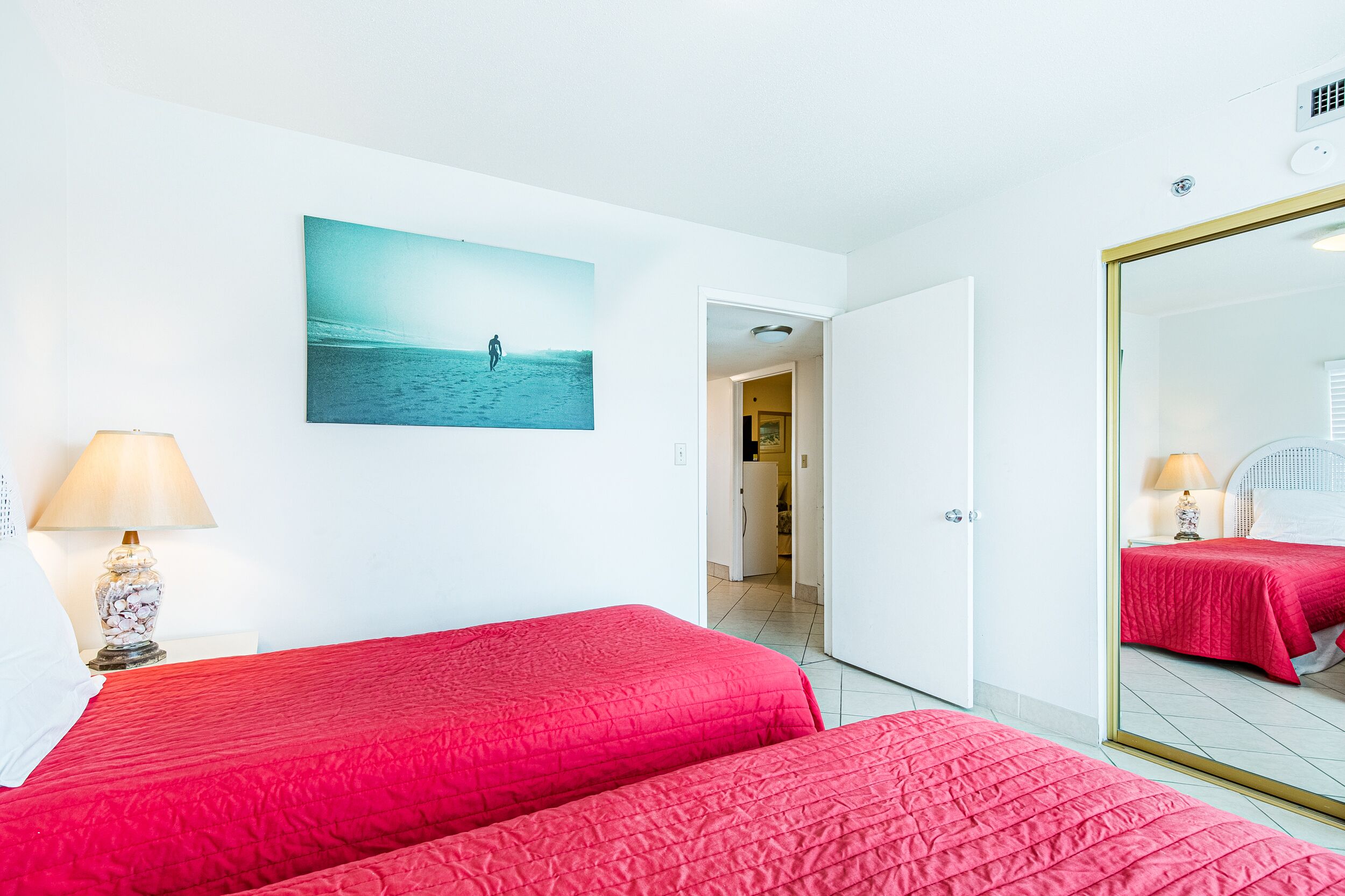 Ein Hotelzimmer mit zwei Betten, einem großen Spiegel, einem Gemälde einer Strandansicht und einer Tür zu einem weiteren Zimmer.
