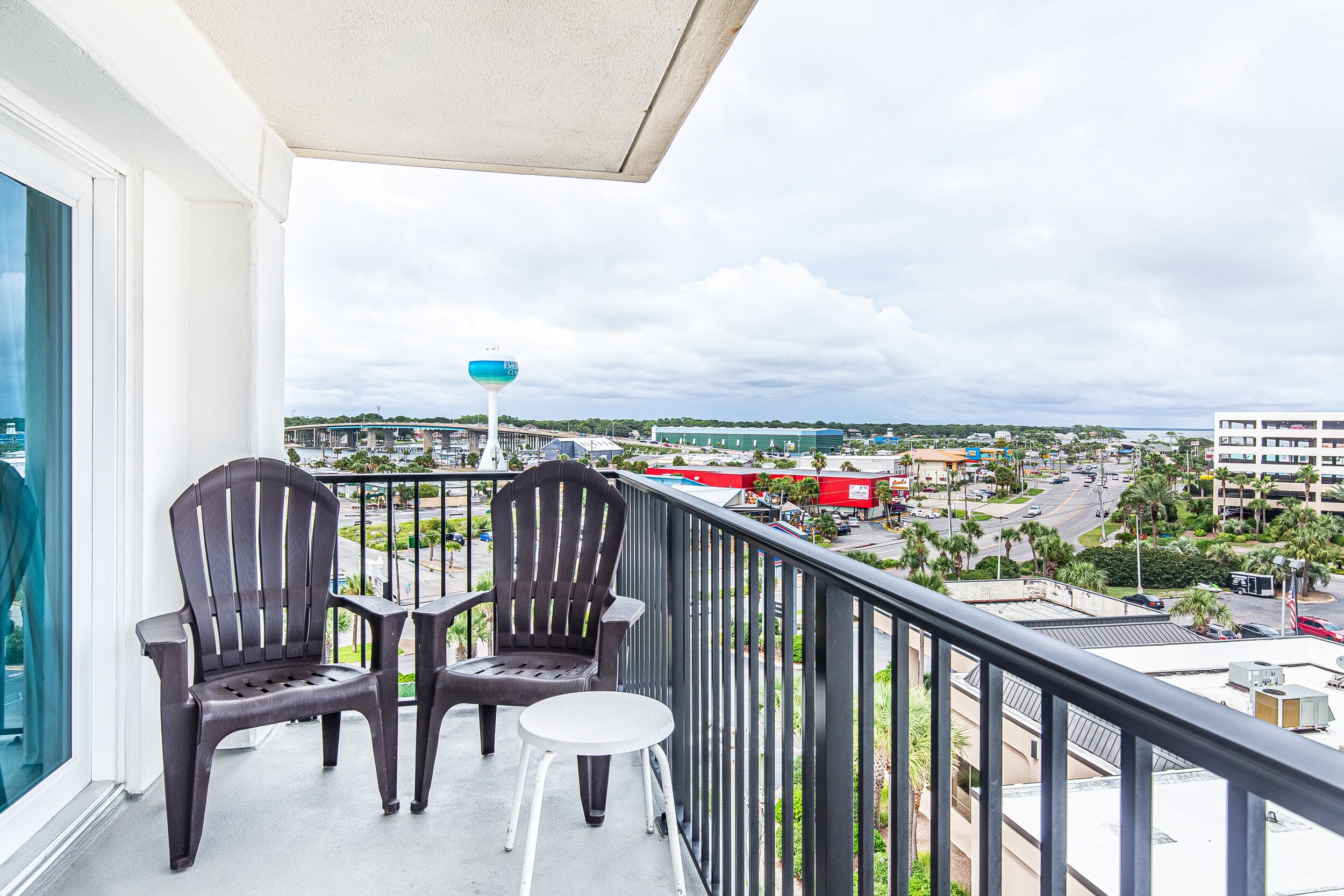 A balcony with two Adirondack chairs and a small table, overlooking a cityscape with buildings and palm trees.