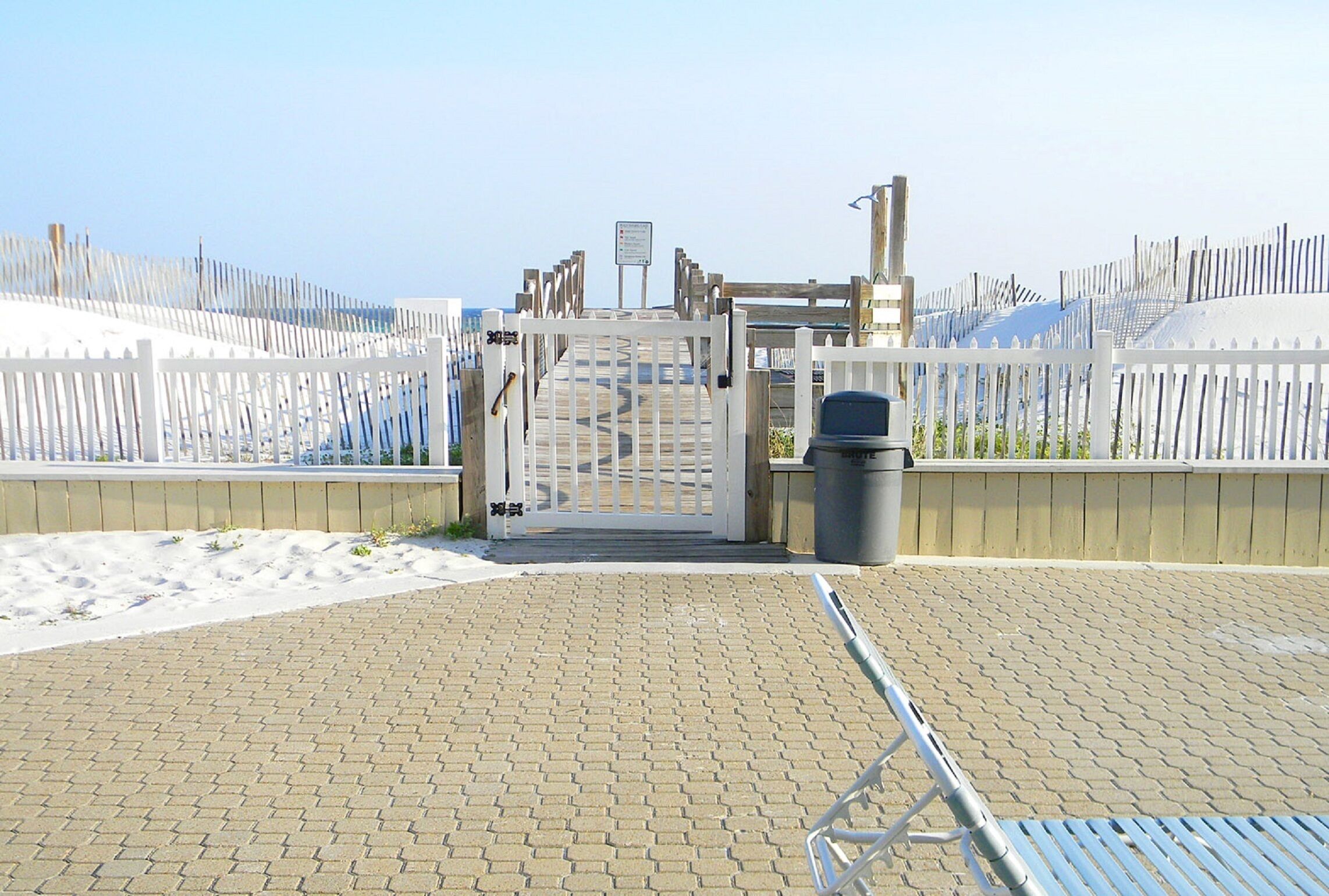 Ein verschlossenes Eingangstor mit einem Metallgitter, einem Mülleimer und einem gepflasterten Weg, der zu einem Strandbereich führt.