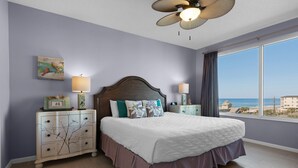 Un dormitorio con una cama grande, dos mesitas de noche, un ventilador de techo, una ventana con vista a la playa y una pared con un cuadro.