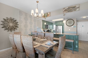 Uma sala de jantar com mesa de madeira e cadeiras de vime, um lustre e uma cozinha ao fundo.