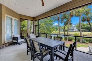 Une terrasse extérieure couverte, dotée d’une table et de chaises, d’un coin salon avec un canapé, et offrant une vue sur un jardin aménagé et un plan d’eau.